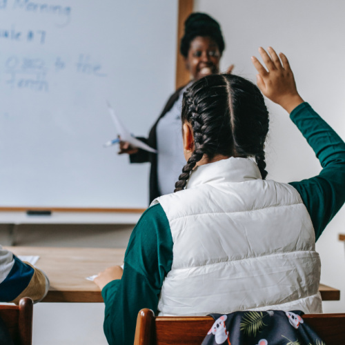 child in classroom with hand raised and teacher in the background