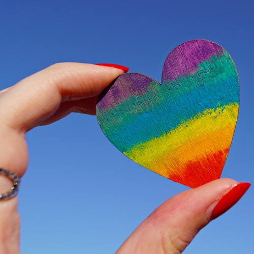 Rainbow-coloured heart being held between a thumb and finger against a blue background.